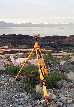 photo of a survey device in rocks; in the distance is seawater, and beyond it, mountains.