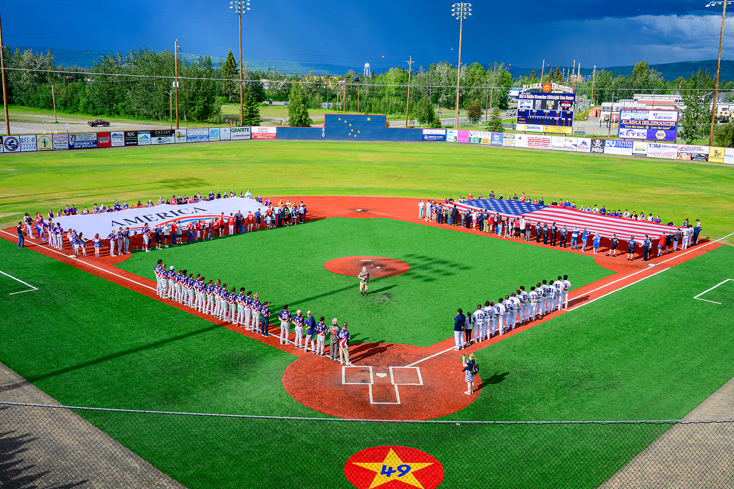 America250-Alaska’s Week of Dreams Closing Ceremony in Fairbanks, July 4, 2026. Players and various people stand on baseball diamond stretching a large american flag on one side and an 'America250' flag on the opposing side.