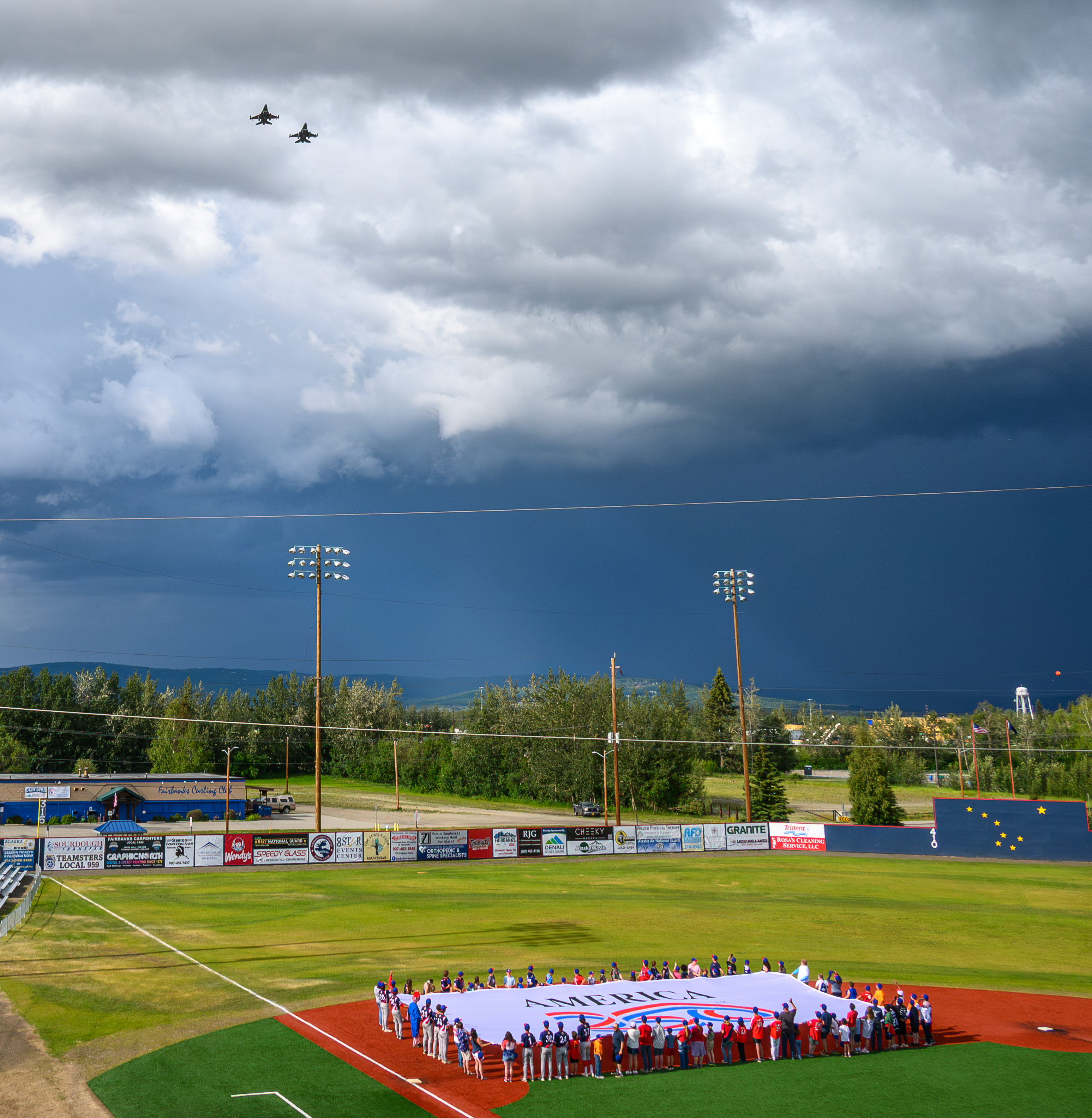 F-18E Super Hornet Fighter Jets fly over baseball field above people stretching out a large America250 flag